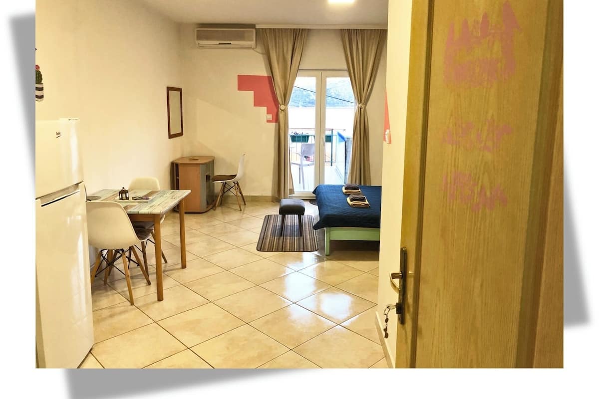 A small, well-lit room with a bed, a desk, a chair, and a refrigerator.