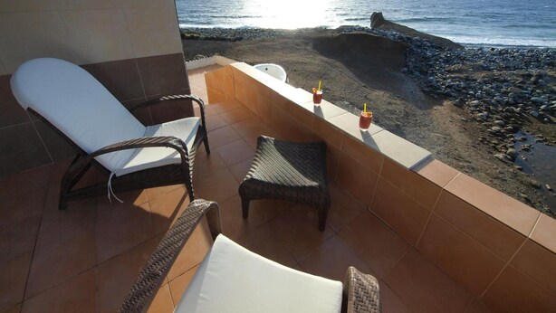 A balcony with wicker chairs, a small table, and a view of the beach and ocean. - Vacation Home 'Sunrise Directly by the Sea' with Sea View, Private Terrace and Wi-Fi (La Mareta)