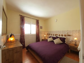 A bedroom with a bed, bedside lamps, a nightstand, a dresser, a window with curtains, and a wall-mounted light fixture.
