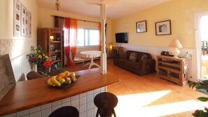 A living room with a sofa, a dining table with fruit, a TV, and a bookshelf. - Vacation Home 'Sunrise Directly by the Sea' with Sea View, Private Terrace and Wi-Fi (La Mareta)