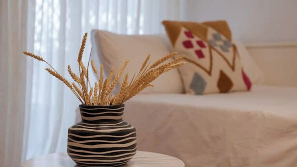 Une pièce avec une table ronde, un vase à rayures zébrées contenant des tiges de blé, et un lit recouvert d’une couverture blanche et agrémenté d’oreillers décoratifs.