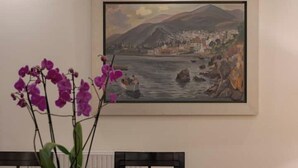 Une peinture encadrée représentant un paysage d’une ville côtière avec des montagnes, une table sur laquelle est posée une orchidée en pot, et une chaise.