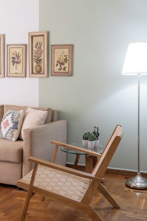 A living room with a sofa, a wooden chair, a glass table with potted plants, and framed floral artwork on the wall.