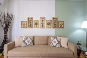 A beige sofa with patterned cushions, a wall with framed floral artwork, and a tall vase of branches. - Best view of Acropolis - Modern 2BR Apt by Meros (Athina)