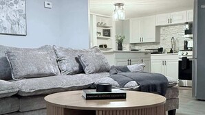 Ein modernes Wohnzimmer mit einem grauen Sofa, einem runden Couchtisch und einer Küche im Hintergrund.