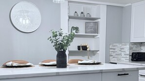 Eine moderne Küche mit weißen Schränken, einer zentralen Kochinsel mit Pflanze, einer Uhr und Pendelleuchten.