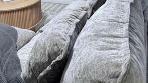 Ein Wohnzimmer mit grauem Sofa, einem runden Holztisch, einem Spiegel an der Wand und einem Fenster mit Jalousien.