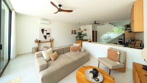 A modern living room with a sofa, a coffee table, and a chair. There is a ceiling fan, a kitchen area with a counter, and a staircase leading to another floor. - Elements Villa A2 - Dreamland (South Kuta)