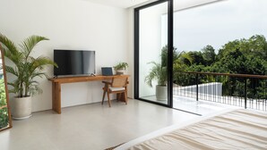 A modern bedroom with a large bed, a wooden desk, a TV, and a balcony with a view of greenery.