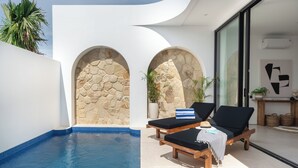 A modern pool area with two lounge chairs, a stone wall, and a glass door leading to a room. - Elements Villa A2 - Dreamland (South Kuta)