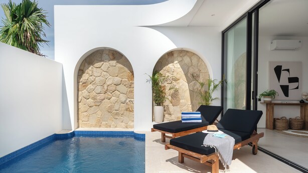 A modern pool area with two lounge chairs, a stone wall, and a glass door leading to a room.