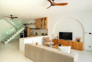 A modern living room with a sofa, TV, and wooden cabinets. There's a staircase with glass railings and a ceiling fan. - Elements Villa A2 - Dreamland (South Kuta)