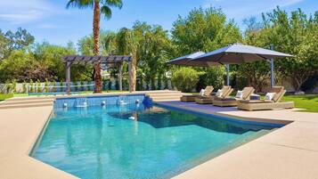 A rectangular swimming pool with clear blue water, surrounded by a paved deck. There are lounge chairs with cushions and a pergola structure.
