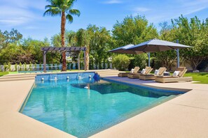 A rectangular swimming pool with clear blue water, surrounded by a paved deck. There are lounge chairs with cushions and a pergola structure.