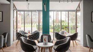 A modern indoor space with a large window, a teal pillar, and a seating area with round tables and chairs. - Holiday Inn NORTHAMPTON by IHG (Northampton)