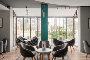 Un espacio interior moderno con un ventanal grande, un pilar verde azulado y una zona de estar con mesas y sillas redondas.
