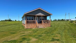 A small, single-story house with a front porch and a red roof, surrounded by a grassy field. - Water Front Cottage- 10 Min Walk To Barachois Beach, Minutes to North Rustico (North Rustico)