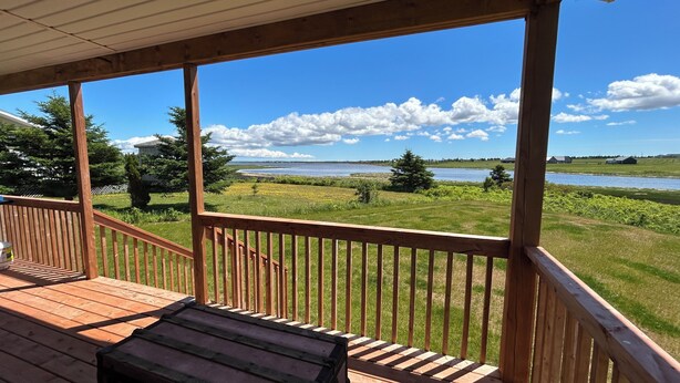 A wooden deck with a bench, overlooking a grassy field and a body of water. - Water Front Cottage- 10 Min Walk To Barachois Beach, Minutes to North Rustico (North Rustico)