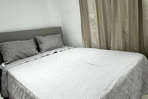 Een bed met een deken met geometrisch patroon, een grijze hoofdbord en een raam met gordijnen.