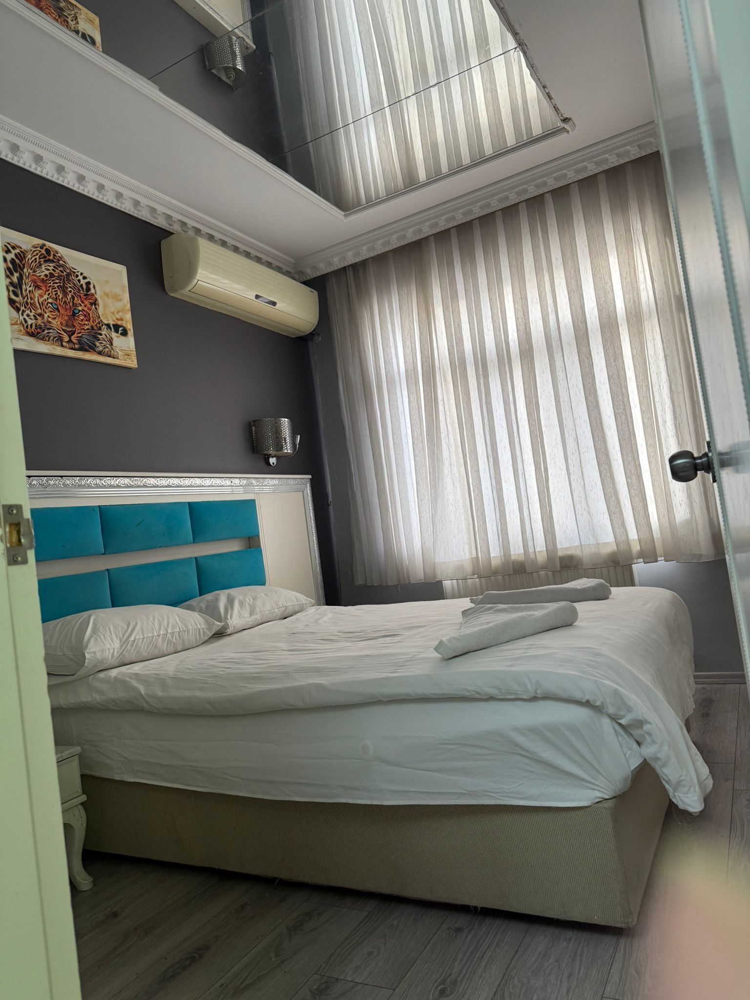 A bedroom with a bed, a headboard, a wall-mounted air conditioner, a window with sheer curtains, and a door.
