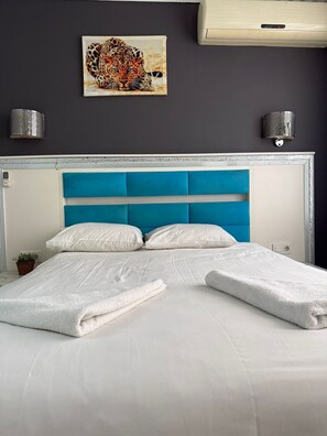 A neatly made bed with white linens and a blue headboard, a wall-mounted artwork of a leopard, and two wall sconces. - HOLİDAY SUİT (İstanbul)