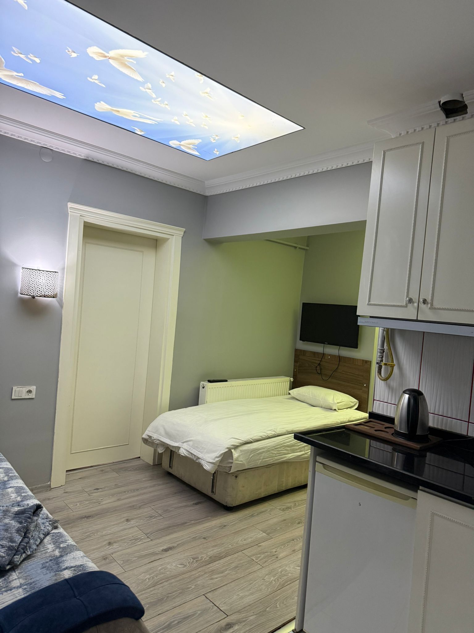 A compact room with a bed, a small kitchenette, a ceiling skylight, and a television.