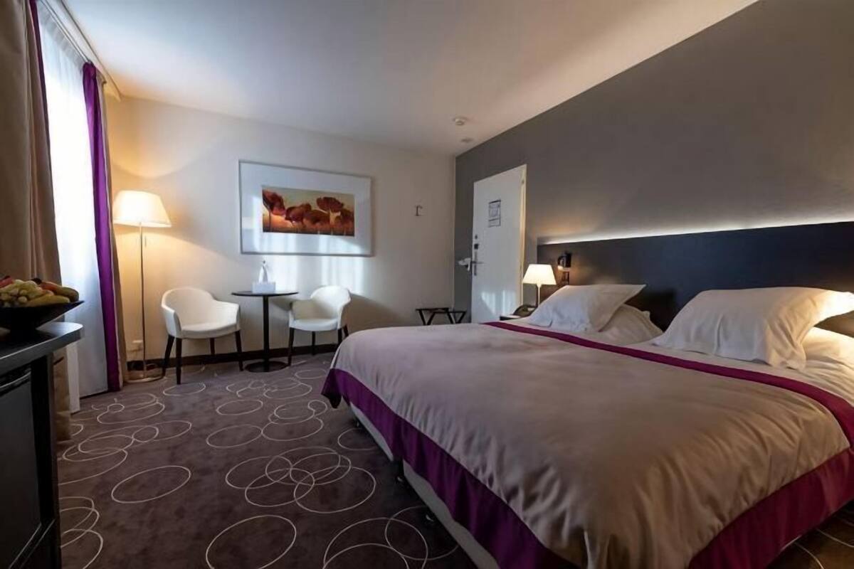 Une chambre d’hôtel avec un lit, deux chaises, un bureau, une lampe, un tableau et une porte.