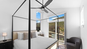 Ein Schlafzimmer mit Himmelbett, Deckenventilator, Nachttisch mit Lampe und Blick auf einen Balkon mit Gartenmöbeln.