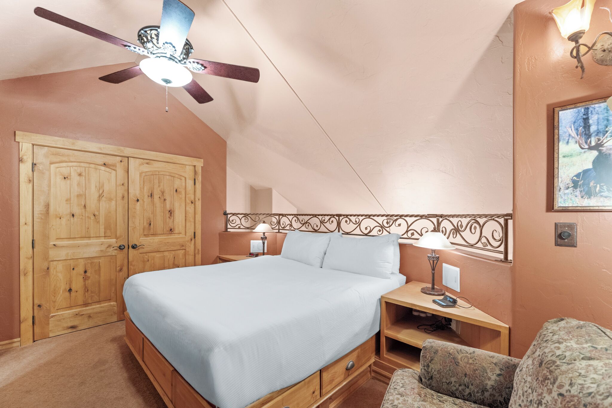 A bedroom with a bed, wooden wardrobe, ceiling fan, bedside lamps, and a framed picture on the wall.