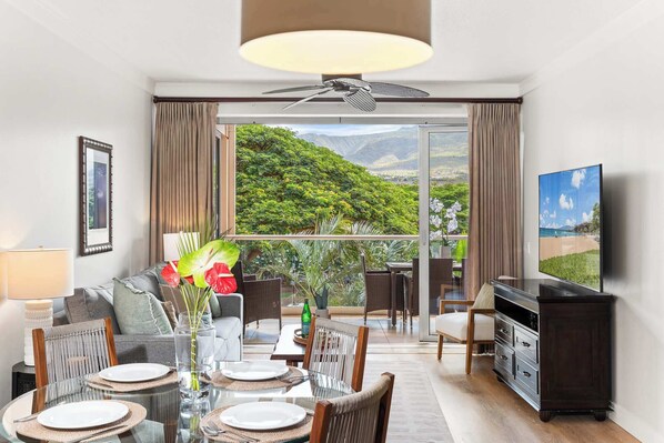 Un salon moderne avec un coin repas, de grandes fenêtres offrant une vue sur les montagnes, un ventilateur de plafond et un téléviseur à écran plat.