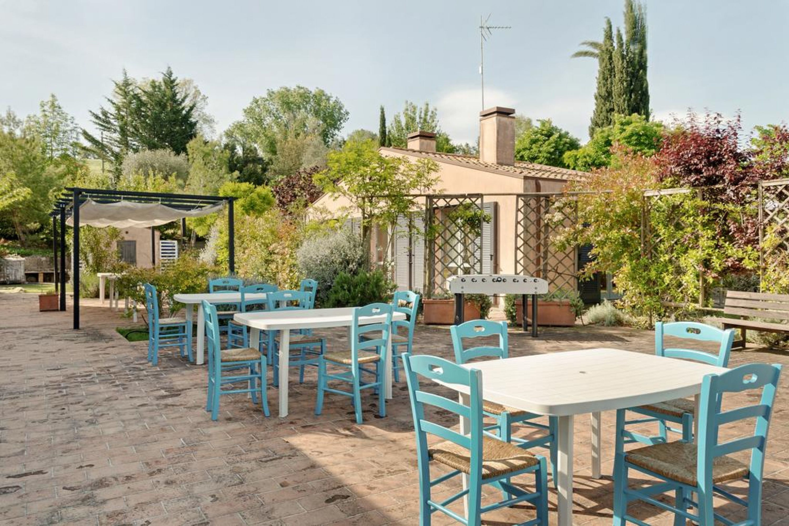 An outdoor patio with blue chairs and white tables, surrounded by greenery and a pergola.