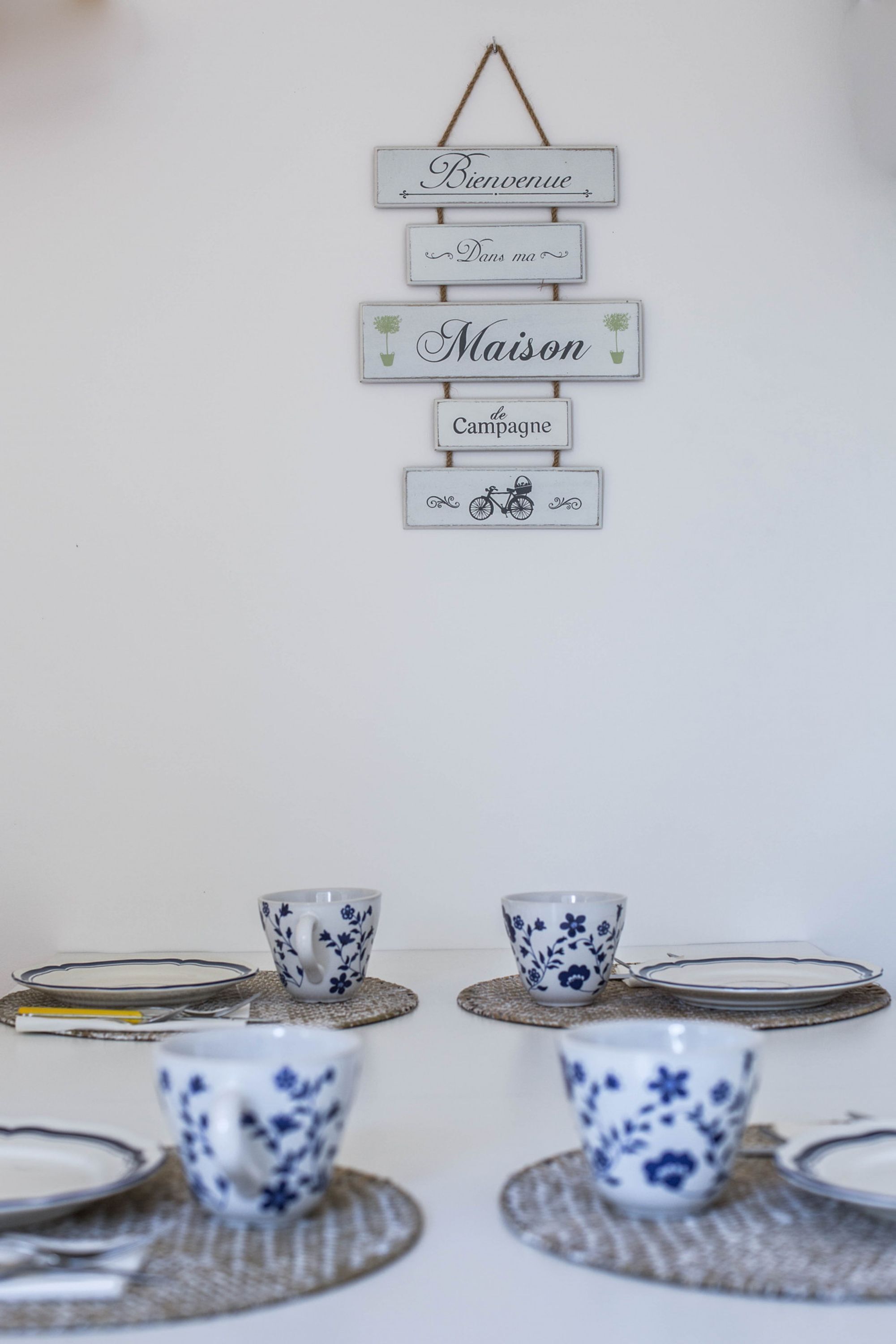 Un letrero en la pared con texto en francés, una mesa con tazas y platitos de estampado floral azul, e una ilustración de una bicicleta.