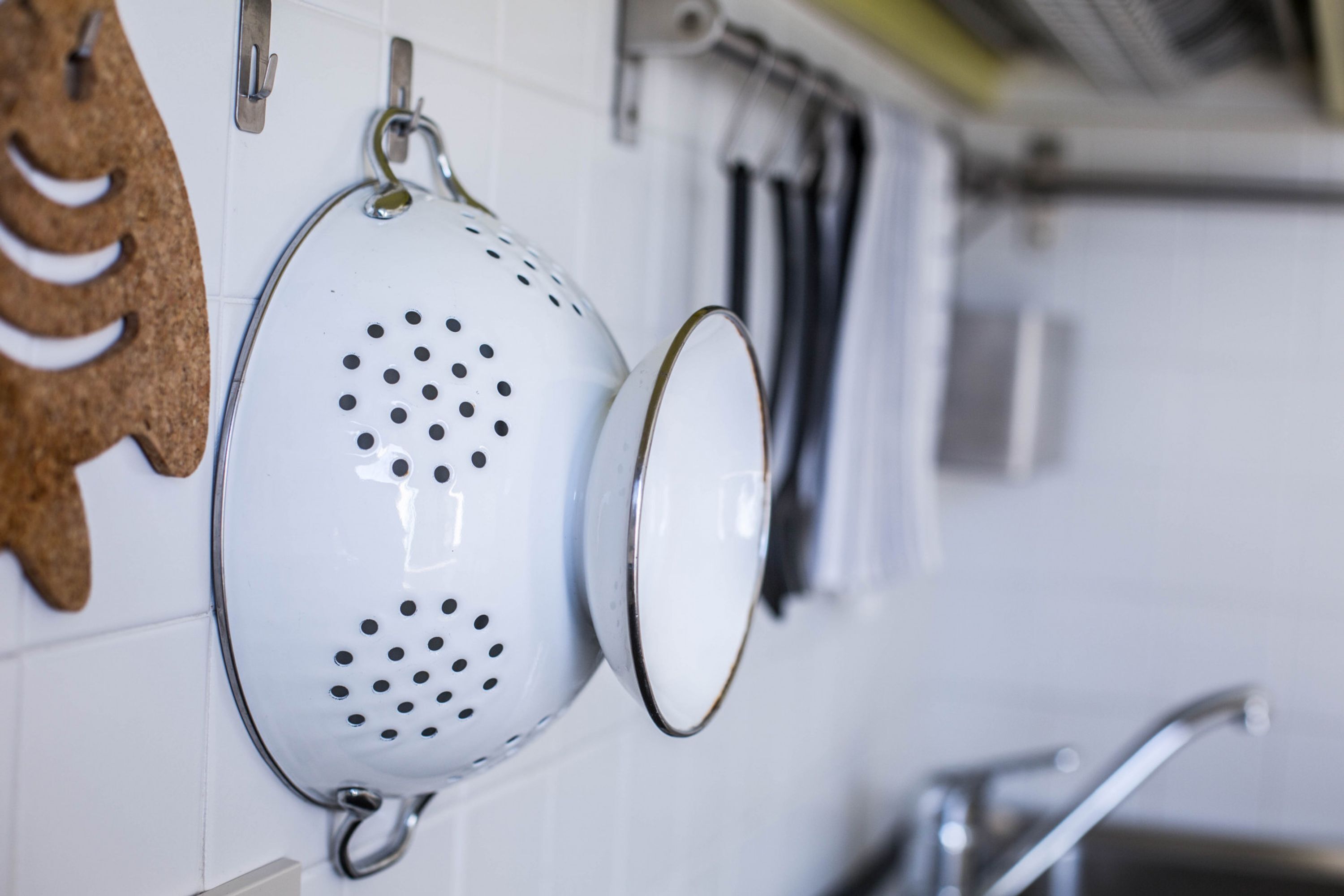 A kitchen with white tiled walls, a hanging colander, and a faucet.