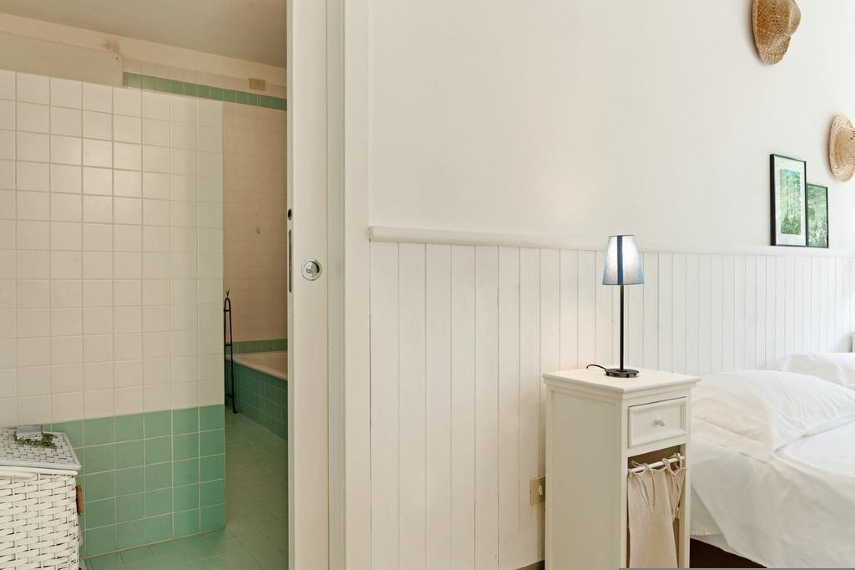 Ein Badezimmer mit weiß-grünen Fliesen, einem weißen Heizkörper, einem Nachttisch mit Lampe und einem Bett mit weißer Bettwäsche.