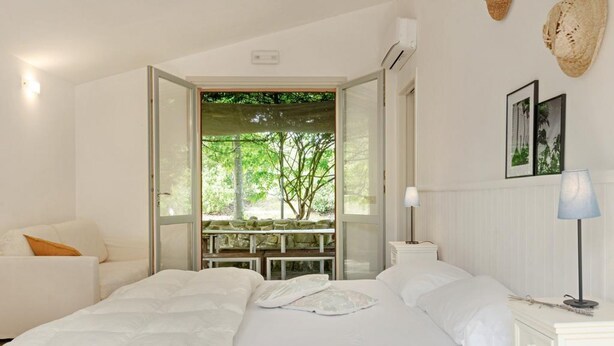 A bedroom with two beds, a white sofa, a table with chairs, and a view of greenery through an open door. - Villa Glicini - Delightful Cottage With Pool (Ostra)