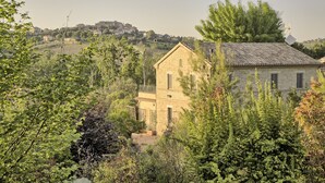 Edificio de piedra con balcón, rodeado de vegetación y con vistas a una colina con casas.