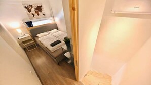 Un couloir étroit mène à une chambre à coucher qui contient un lit, une table de chevet, une lampe et une petite plante.