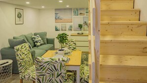 A modern living room with a wooden staircase, a green sofa, patterned chairs, and a dining table with a potted plant. - Cosy flat in Akácfa str., Aya (Budapest)