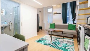 A compact living space with a kitchenette, a sofa with green cushions, a small table, and a glass shower enclosure.