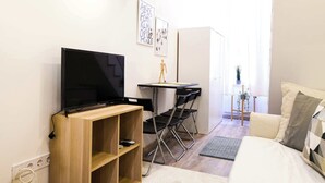 Ein modernes, gut beleuchtetes Zimmer mit einem Flachbildfernseher auf einem Holzständer, einem Schreibtisch mit Stuhl, einer Couch und einem Korbstuhl.