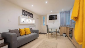 Una sala de estar moderna con un sofá gris, cojines amarillos, una mesa de comedor con sillas, un televisor montado en la pared y una zona de cocina con armarios y un fregadero.