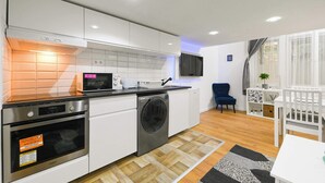 A modern kitchen with white cabinets, a microwave, an oven, and a washing machine. There is a dining area with a table and chairs, and a blue chair.