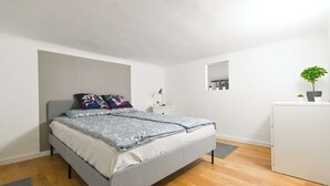 A modern bedroom with a grey bed, white walls, a small bedside table, a mirror, and a potted plant.