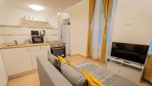 A compact living space with a kitchenette, a sofa, a television, and a window with curtains. - Klauzal  · 2 bedroom downtown flat in Budapest, Lisa (Budapest)