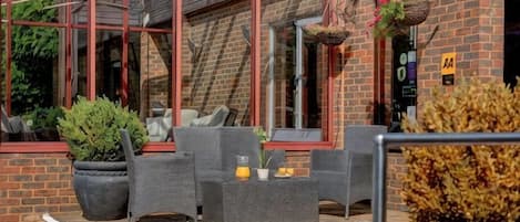 An outdoor seating area with wicker furniture, potted plants, and a brick building with a red frame.