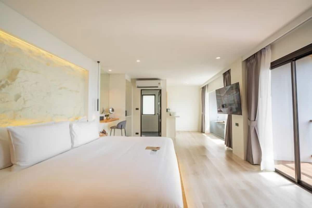 A modern hotel room with a large bed, a desk, a TV, and a view through the glass door.