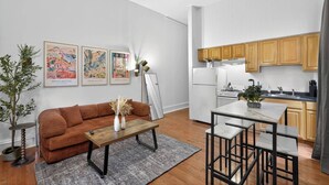 A modern living room with a brown sofa, a coffee table, and a dining area with a white table and black chairs. There are three framed artworks on the wall, a potted plant, and a kitchen area with wooden cabinets and a stove. - 1/1 Center City Sleeps 6 Fast Wifi (Philadelphia)