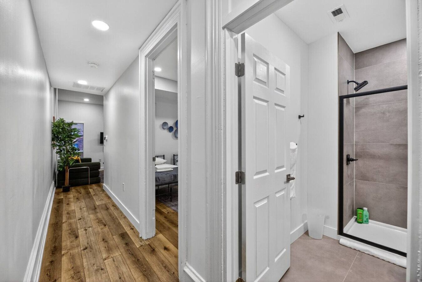 A hallway with wooden flooring, white walls, and a door leading to a bathroom with a shower and tiled walls.