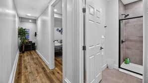 Un pasillo con piso de madera, paredes blancas y una puerta que da a un baño con ducha y paredes de azulejos.