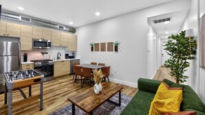 Una cocina moderna con armarios de madera, un comedor con mesa y sillas, una mesa de futbolín y un sofá verde con un cojín amarillo.